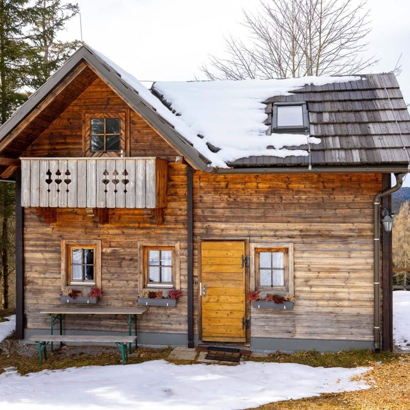 Gemütliches Holzhaus im Schnee mit Balkon und blühenden Fensterkästen, umgeben von einer winterlichen Landschaft. Ideal für eine romantische Auszeit oder einen Familienurlaub in der Natur.