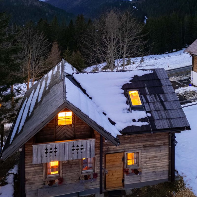 Holzhaus in den Bergen mit schneebedecktem Dach und beleuchteten Fenstern bei Sonnenuntergang. Die Umgebung zeigt eine beeindruckende Berglandschaft mit Wäldern und schneebedeckten Hügeln. Ideal für eine gemütliche Auszeit in der Natur.