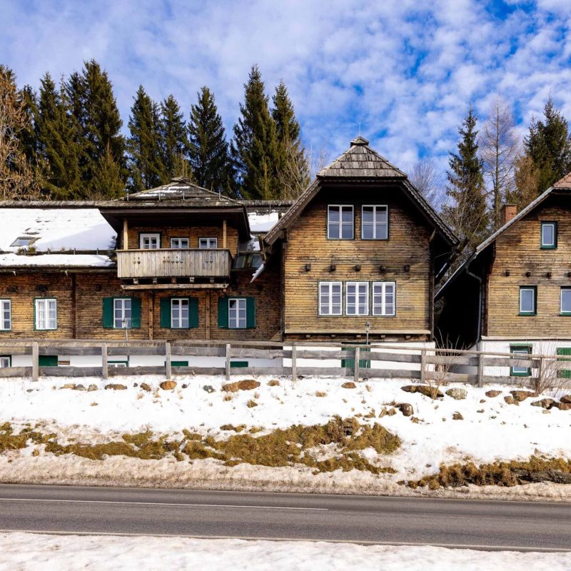 Altes Holzhaus im alpinen Stil mit Schnee auf dem Dach, umgeben von Nadelbäumen und einer Straße im Vordergrund. Die Fassade zeigt mehrere Fenster mit grünen Rahmen und einen Balkon.