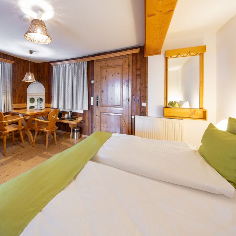 Gemütliches Zimmer mit Doppelbett und grünen Kissen, Holzverkleidung, Essbereich mit Holztisch und Stühlen, sowie Fenster mit Vorhängen. Ideal für einen entspannten Aufenthalt in der Natur.