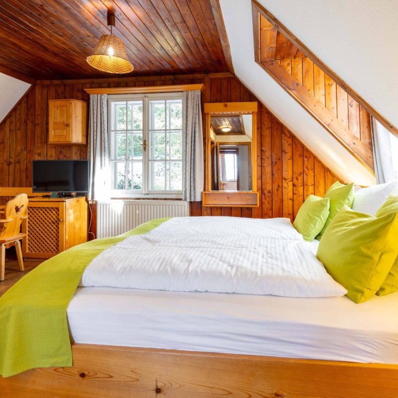 Gemütliches Schlafzimmer mit Holzvertäfelung, einem großen Bett mit weißen Bettwaren und grünen Kissen, einem Schreibtisch, einem Fernseher und einem Fenster, das Tageslicht hereinlässt. Ideal für einen entspannten Aufenthalt in einer rustikalen Umgebung.
