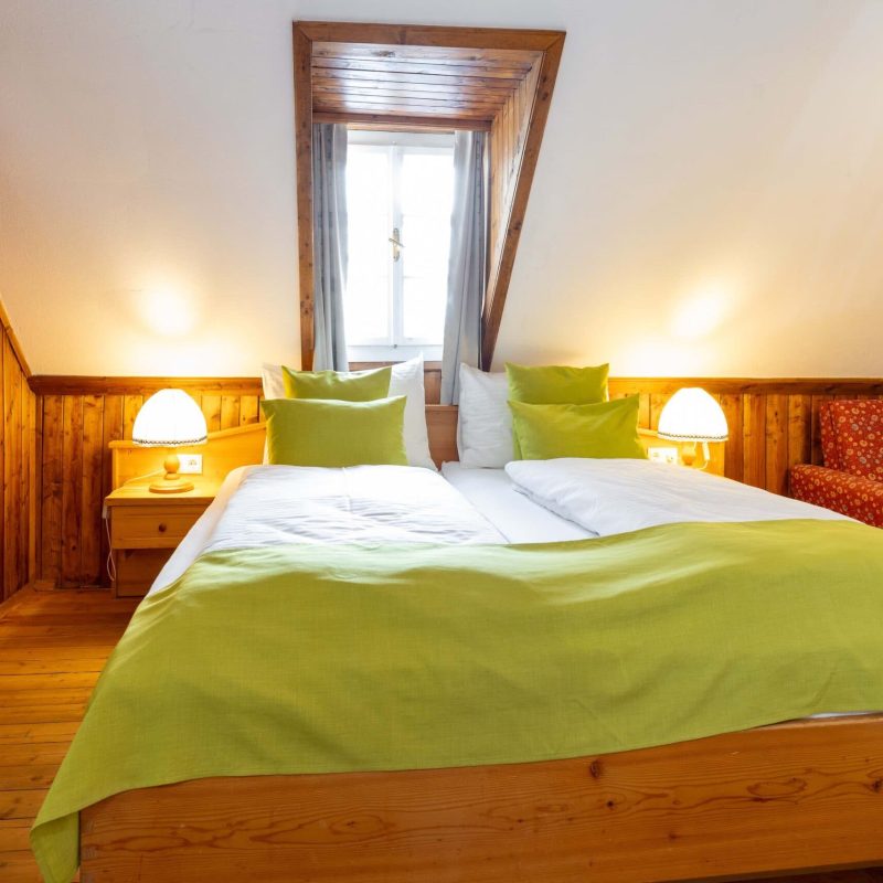 Gemütliches Schlafzimmer mit einem großen Bett, grünem Bettzeug und hellen Kissen, umgeben von Holzverkleidungen. Neben dem Bett steht ein Nachttisch mit einer Lampe, und ein roter Sessel ergänzt die Einrichtung. Fenster sorgt für Tageslicht und eine einladende Atmosphäre. Ideal für einen entspannten Aufenthalt.