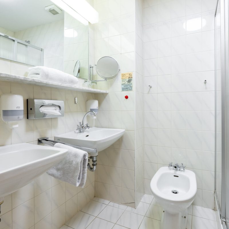 Modernes Badezimmer mit zwei Waschbecken, einem Bidet und einer Duschkabine. Die Wände sind hell gefliest, und eine große Spiegeloberfläche sorgt für eine helle Atmosphäre. Handtücher und Seifenspender sind gut sichtbar, was auf eine gepflegte Hygiene hinweist.