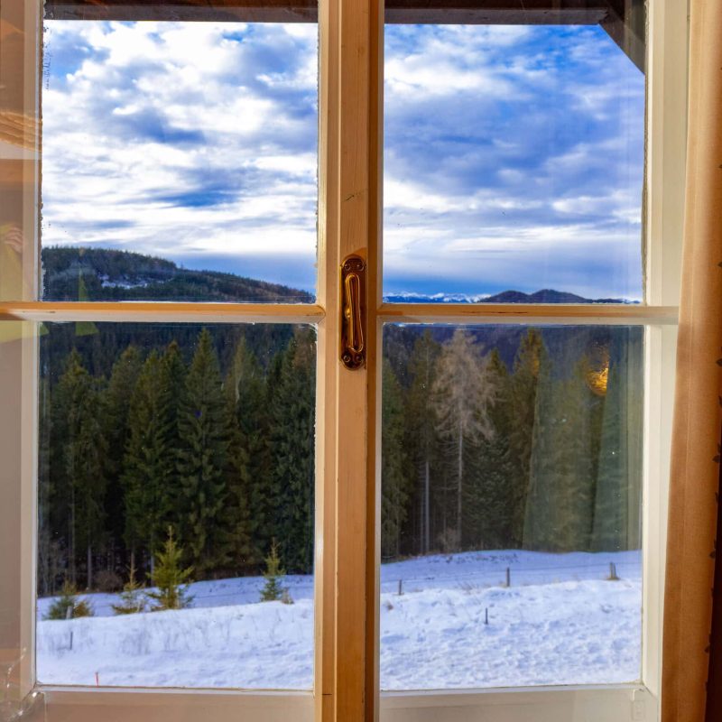 Blick aus einem Fenster auf eine verschneite Landschaft mit Nadelbäumen und Bergen im Hintergrund, umrahmt von einem Vorhang in sanften Farben. Ideal für winterliche Urlaubs- oder Naturthemen.