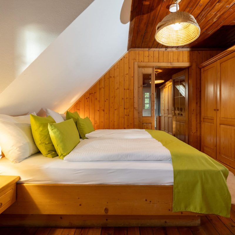 Gemütliches Schlafzimmer mit Holzvertäfelung, einem Doppelbett mit weißen Bettlaken und grünen Kissen, sowie einer warmen Nachttischlampe. Der Raum ist hell erleuchtet und bietet eine einladende Atmosphäre. Im Hintergrund sind Holzschränke sichtbar, die zusätzlichen Stauraum bieten. Ideal für einen entspannten Aufenthalt.