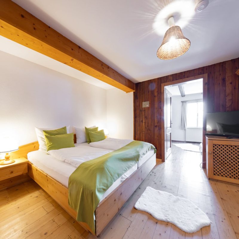 Gemütliches Schlafzimmer mit einem Doppelbett, ausgestattet mit grünen Kissen und einer weißen Bettdecke. Der Raum verfügt über einen Holzboden, eine Nachttischlampe und einen Fernseher. Holzverkleidungen an der Wand und eine offene Tür zu einem weiteren Raum verleihen dem Interieur eine warme Atmosphäre.