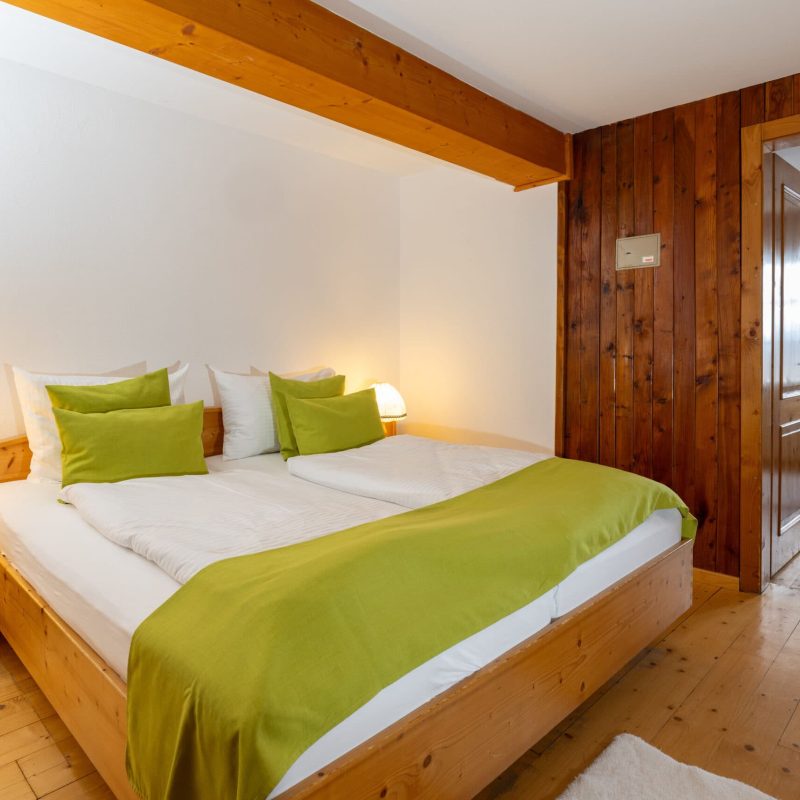 Gemütliches Schlafzimmer mit einem Doppelbett, grünen Kissen und einer warmen Tischlampe, umgeben von Holzvertäfelungen und einem offenen Türrahmen, der in einen weiteren Raum führt. Ideal für einen entspannten Aufenthalt.