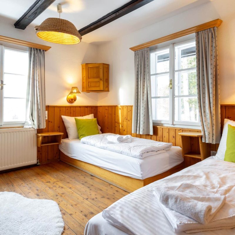 Helles und gemütliches Schlafzimmer mit zwei Einzelbetten, Holzverkleidung und großen Fenstern. Die Einrichtung umfasst eine grüne Kissen und eine stilvolle Lampe. Ideal für entspannte Übernachtungen in einer ruhigen Umgebung.