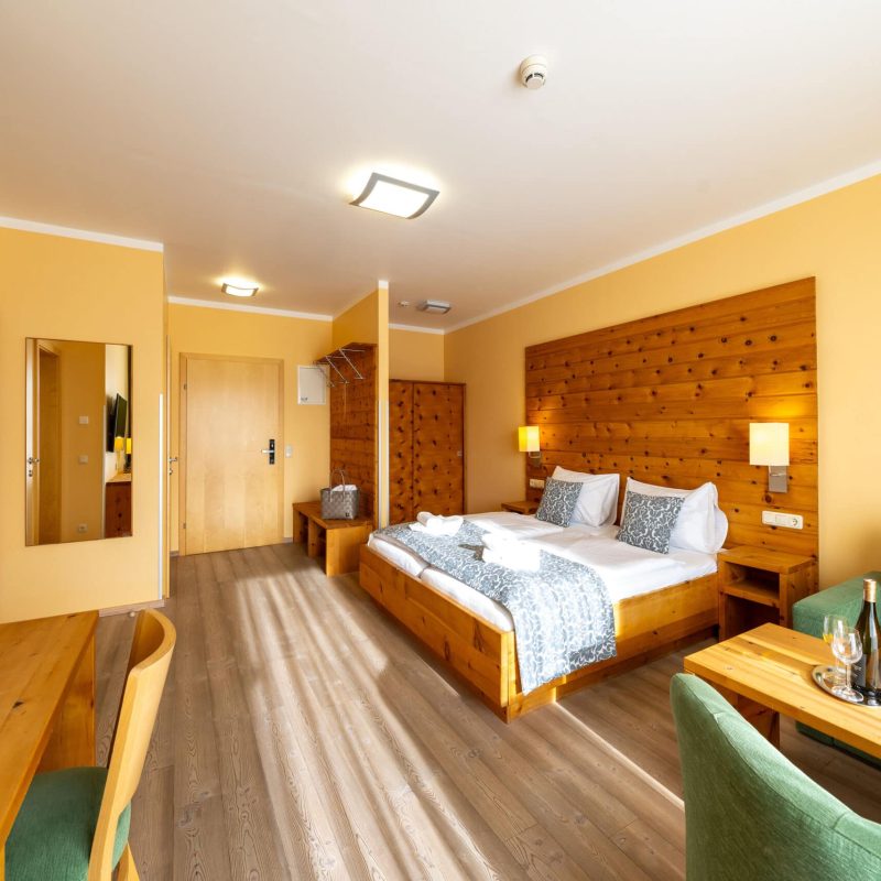 Modernes Hotelzimmer mit Holzmöbeln, Doppelbett, gemütlicher Sitzecke und Schreibtisch. Das Zimmer bietet eine warme Atmosphäre mit hellen Wänden und natürlichem Licht. Ideal für entspannte Aufenthalte oder Geschäftsreisen.