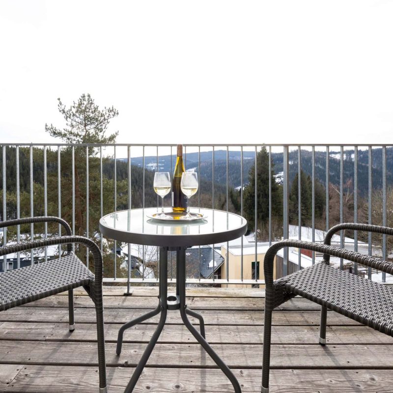 Balkon mit Blick auf die Berge, ausgestattet mit einem runden Tisch und zwei Stühlen, auf dem Tisch stehen zwei Gläser Weißwein und eine Flasche Wein. Ideal für entspannte Momente in der Natur.