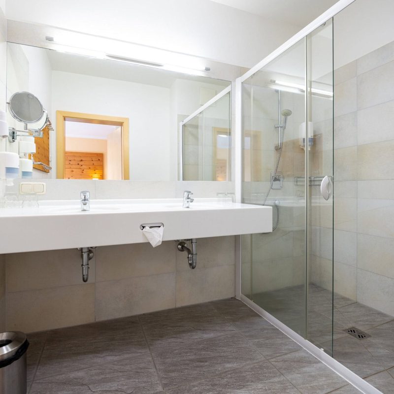 Modernes Badezimmer mit gläserner Dusche, hellen Fliesen und einem großen Waschbecken. Im Hintergrund sind Holzdetails und ein Spiegel sichtbar. Ideal für eine entspannende Auszeit.
