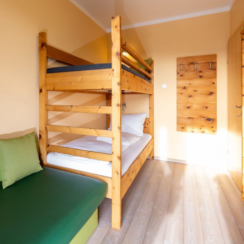 Gemütliches Zimmer mit Etagenbett aus Holz, einem grünen Sofa und natürlichem Licht. Ideal für Familien oder Gruppen, die eine komfortable Unterkunft suchen. Helle Wände und Holzdetails schaffen eine einladende Atmosphäre.