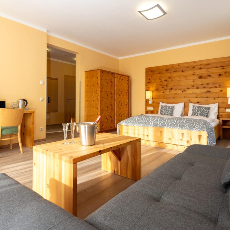 Helles und modernes Hotelzimmer mit Holzdekoren, bestehend aus einem Doppelbett, einem Sofa, einem Couchtisch und einem Arbeitsplatz mit Fernseher. Die warmen Farben und die gemütliche Einrichtung schaffen eine einladende Atmosphäre. Ideal für Erholung und Entspannung während des Aufenthalts.