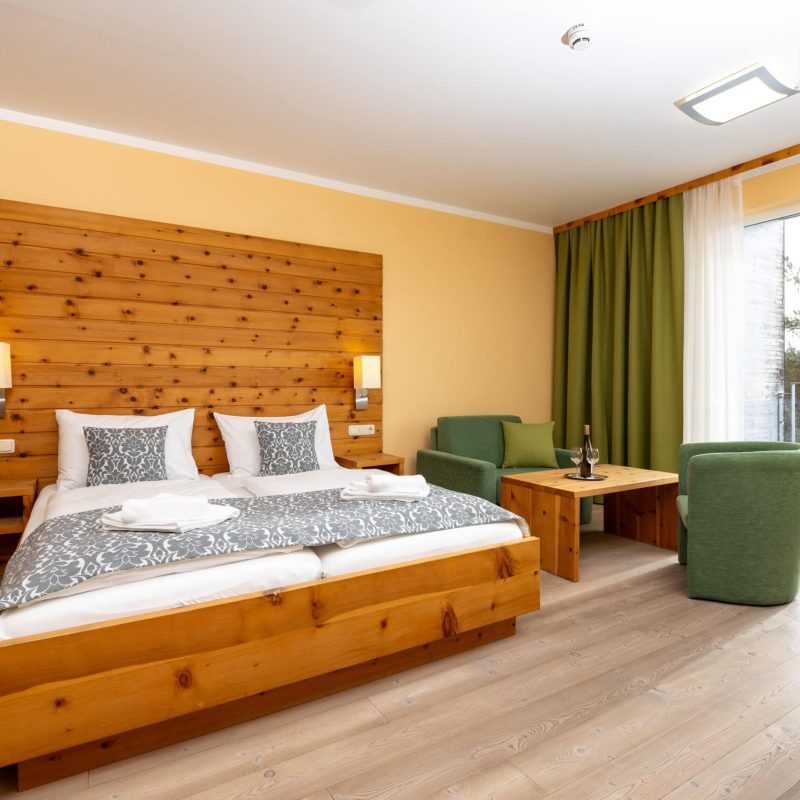 Gemütliches Hotelzimmer mit Doppelbett, Holzverkleidung und modernen Möbeln. Das Zimmer verfügt über eine Sitzecke mit grünen Stühlen und einem Holztisch sowie große Fenster, die viel Tageslicht hereinlassen und einen Blick auf die Natur bieten. Ideal für entspannte Aufenthalte.