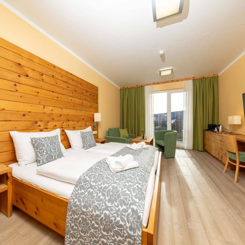 Modernes Hotelzimmer mit Holzwand, Doppelbett, gemütlichen Sesseln und Schreibtisch. Große Fenster mit Vorhängen und Blick auf die Umgebung. Fernseher und Annehmlichkeiten wie Wasserkocher vorhanden. Ideal für Entspannung und Komfort während des Aufenthalts.