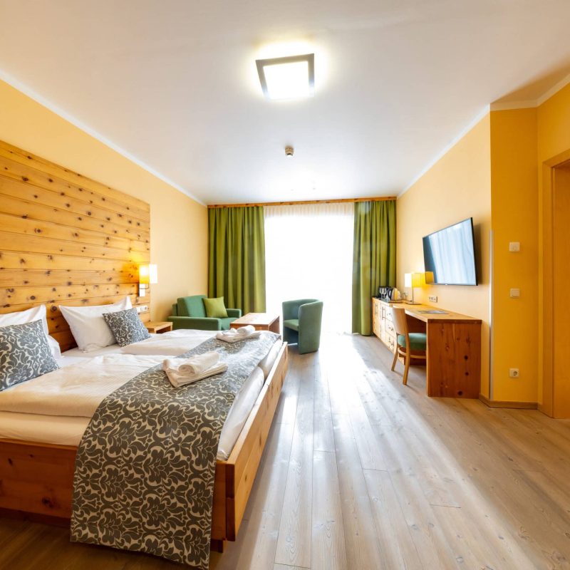 Helles und einladendes Hotelzimmer mit Holzdekor, zwei Einzelbetten, gemütlicher Sitzecke, Schreibtisch, Fernseher und Blick auf einen Balkon. Ideal für Familien oder Paare, die einen komfortablen Aufenthalt suchen.