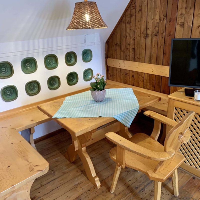 Gemütlicher Essbereich mit holzfarbenem Tisch, Stühlen und einer dekorativen Tischdecke in Blau. Eine Hängeleuchte sorgt für angenehmes Licht, während eine kleine Pflanze in einer Vase den Tisch schmückt. Im Hintergrund sind grüne Wandmalereien zu sehen, die dem Raum eine einladende Atmosphäre verleihen.