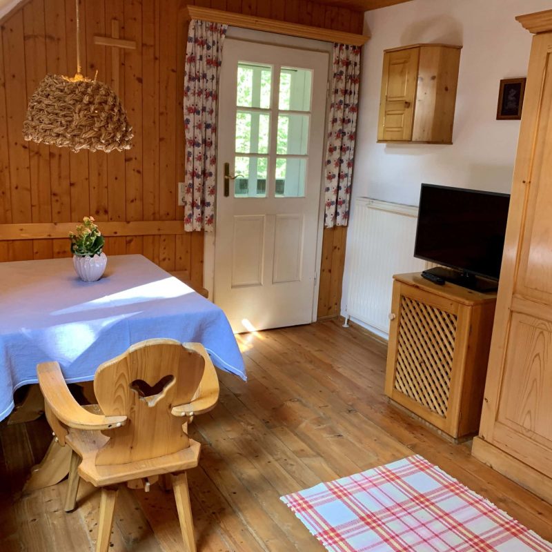 Gemütlicher Wohnraum mit Holzvertäfelung, einem Tisch mit blauer Tischdecke, einem dekorativen Stuhl mit Herzmotiv und einem Fernseher. Die Fenster sind mit blumigen Vorhängen geschmückt und der Boden ist aus Holz. Ein kariertes Teppichstück ergänzt die rustikale Einrichtung. Ideal für einen entspannten Aufenthalt in der Natur.