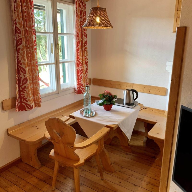 Gemütliche Essecke in einer rustikalen Unterkunft mit Holzbank, Stuhl und Tisch, dekoriert mit einer Tischdecke, einer Vase und einer Pflanze. Helle Fenster mit bunten Vorhängen sorgen für eine freundliche Atmosphäre. Die warmen Holzböden und die Beleuchtung schaffen eine einladende Umgebung.